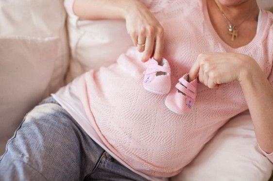 Acercamiento a mujer embarazada con blisa rosa y jeans sostiene unos zapatitos de bébe rosa