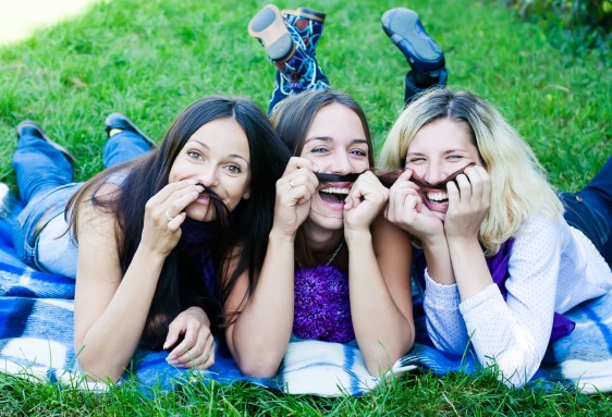 Tres mujeres recostadas en el pasto con su cabello simulando bifotes y sonriendo