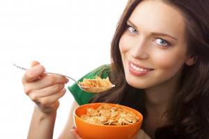 Mujer sonriendo con camisa verde y con una cuchara en la mano, con la otra sosteniendo plato hondo naranja que contiene cereal