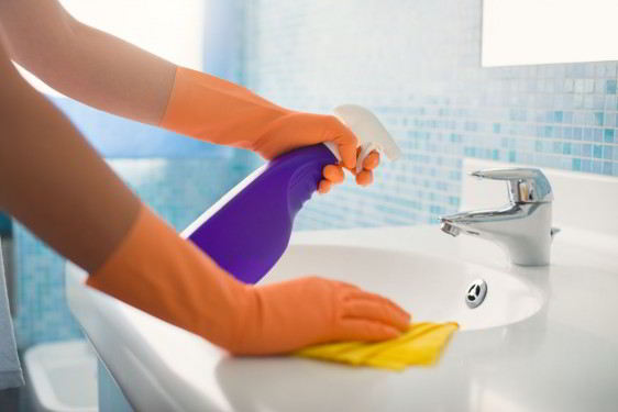 Acercamiento a manos con guantes naranja en un lavamanos limpiando con un trapo y una botella