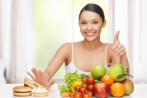 Mujer rechazando con la mano derecha comida rápida enfrente tiene frutas y con la mano izquierda levanta el dedo pulgar