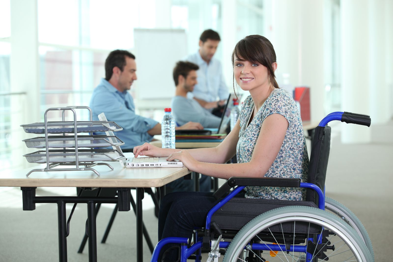 Mujer sentada en silla de ruedas en una oficina trabajando con una computadora al fondo oficinistas trabajando