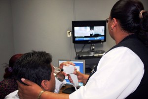 Enfermera con aparato en una mano sentado una persona que observa de cerva el aparato de la enfermera al fondo una pantalla con un instructor