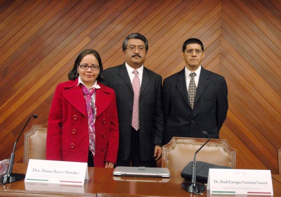 Diana Reyes Morales con saco rojo y Raúl Enrique Guzmán García con Eugenio Vázquez Meraz de pie al fondo una pared de madera