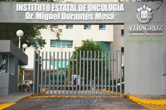 Imagen de la entrada al Instituto Estatal de Oncología de Veracruz, barda gris con el texto de la instutución en la parte superior al fondo un edificio amarillo