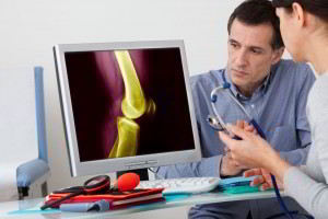 Una doctora observa una pantalla con simulación de huesos el paciente al fondo escucha