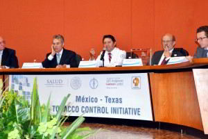 Grupo de personas sentadas en u amesa abajo un letrero con el texto "México - Texas TABACO CONTROL INICIATIVE"
