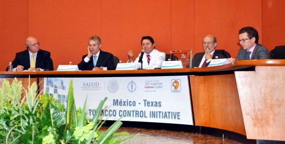 Grupo de personas sentadas en u amesa abajo un letrero con el texto "México - Texas TABACO CONTROL INICIATIVE"
