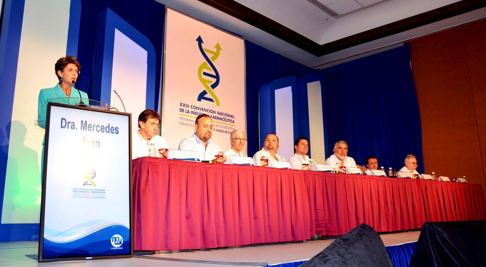 Personas sentadas en una mesa Mercede Juan de pie en un podium atras un letrero "XXIII Convención Nacional de la Industria Farmacéutica"