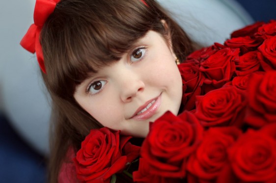 Acercamiento al rostro de una niña con un moño rojo y rosas rojas en sus manos