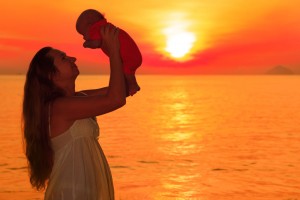 Silueta de mujer cargando un bebé con sus manos al fondo un amanecer naranja