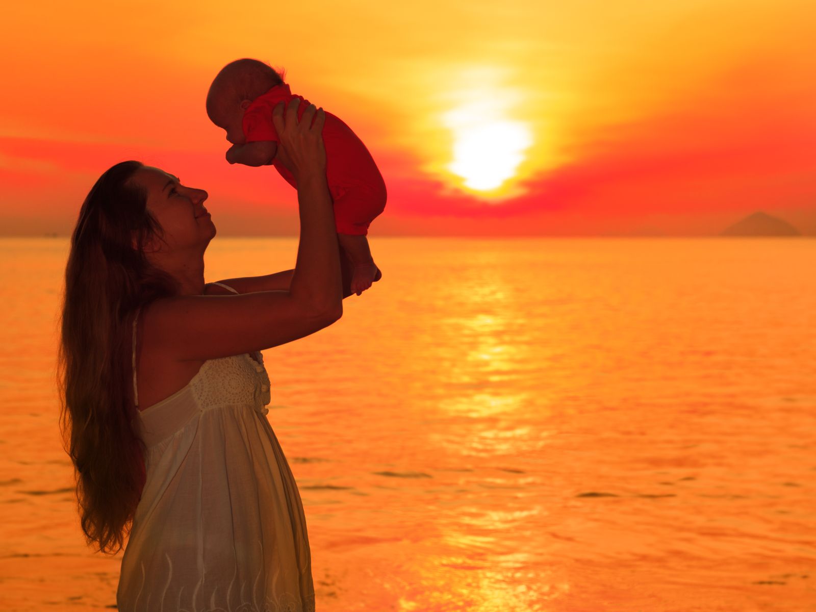 Silueta de mujer cargando un bebé con sus manos al fondo un amanecer naranja