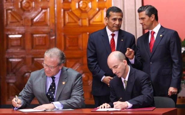 Dos funcionarios firmando dcymentos en una mesa atras de ellos dos presidentes observando