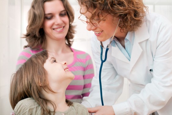 Doctora revisa co estetoscopio a una niña en el fondo una mujer observando
