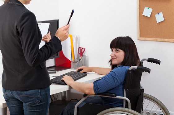 Mujer en silla de ruedas trabajando enfrente de una computadora escucha a una mujer de pie