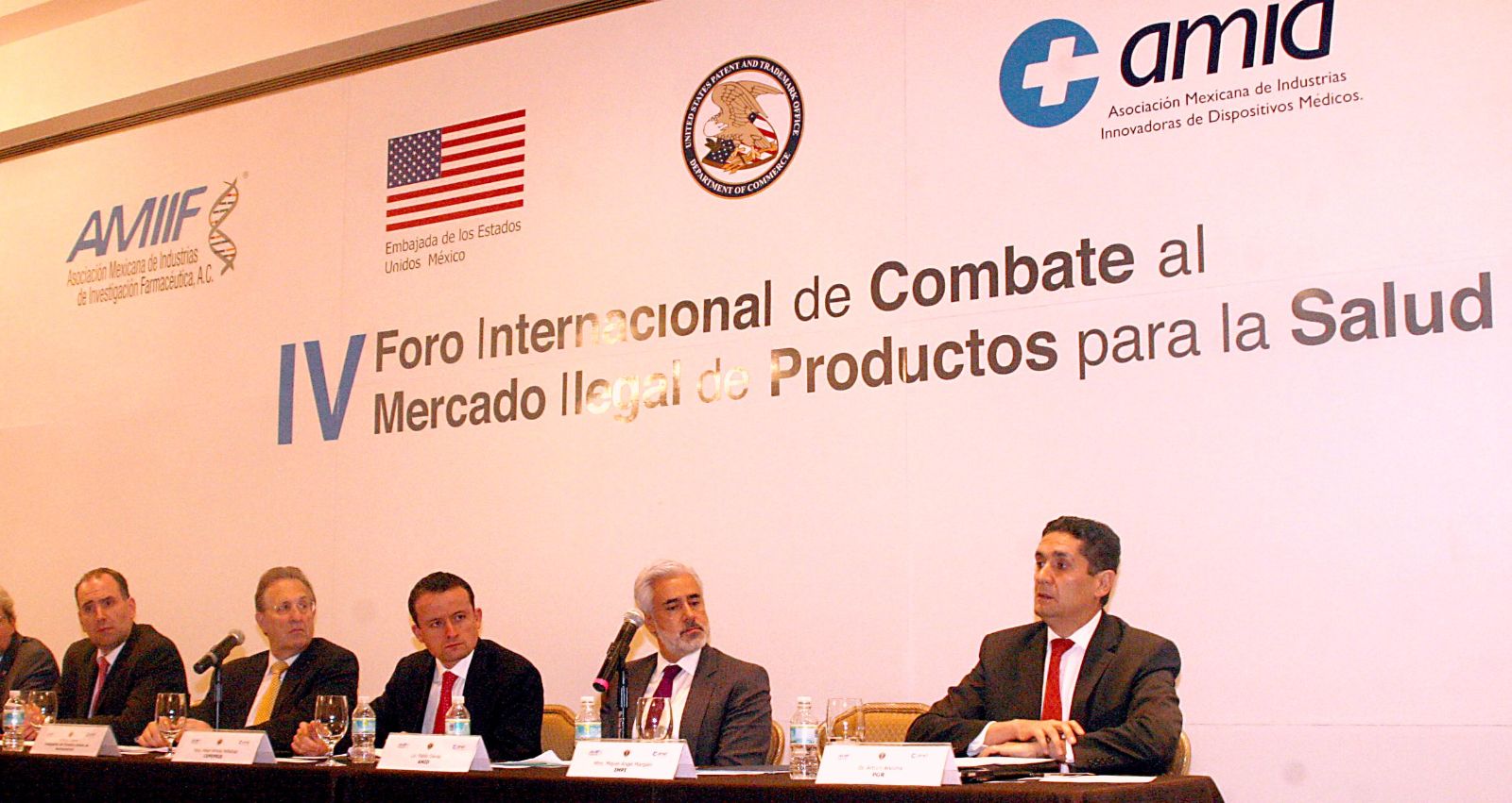 Mikel Arriola, Anthony Wayne, Miguel Ángel Margáin, entre otros sentados en una mesa al fondo un anuncio .del IV Foro Internacional de Combate al Mercado Ilegal de Productos para la Salud