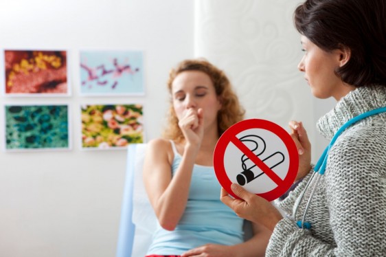 Doctora sosteniendo letrero de prohibido fumar al fondo una paciente acostada tosiendo