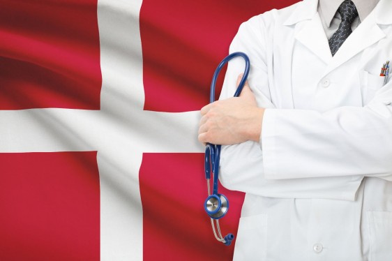 Acercamiento al cuerpo medio de un médico que sostiene un estetoscopio al fondo la bandera de dinamarca