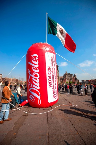 Globo rojo de cinco metros de altura conletras blancas manuscritas con la palabra "Diabetes" en el zócalo de la ciudad de M+exico al fondo la bandera de los Estados Unidos Mexicanos