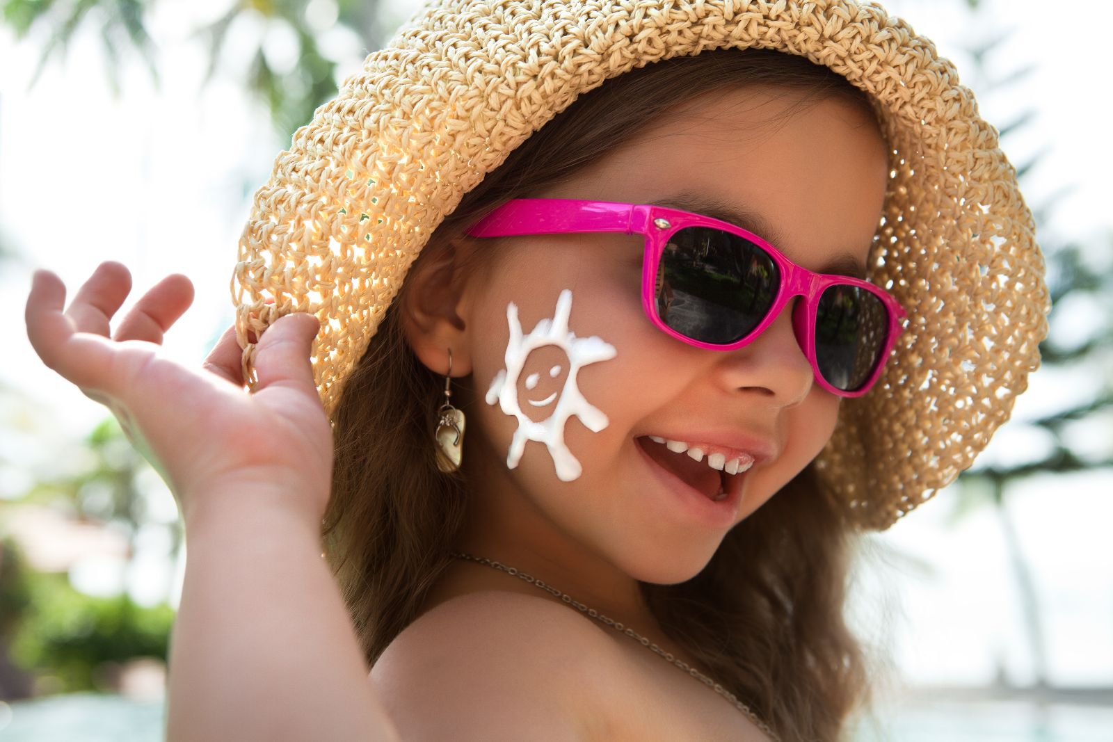 Niña con sombrero y lentes sonriendo con protector solar aplicado en la mejilla con la forma de un sol