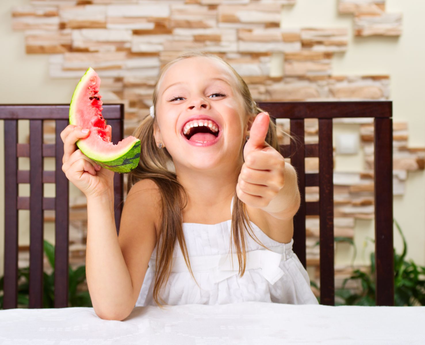 Niña sonriendo con una rebanada de sandía en una mano y en la otra con el pulgar hacia arriba