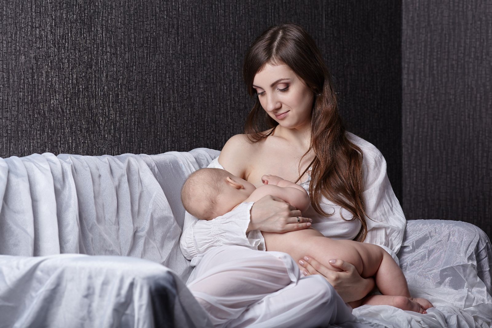 Una madre con vestido blanco en un sofá blanco amamantando a su bebé