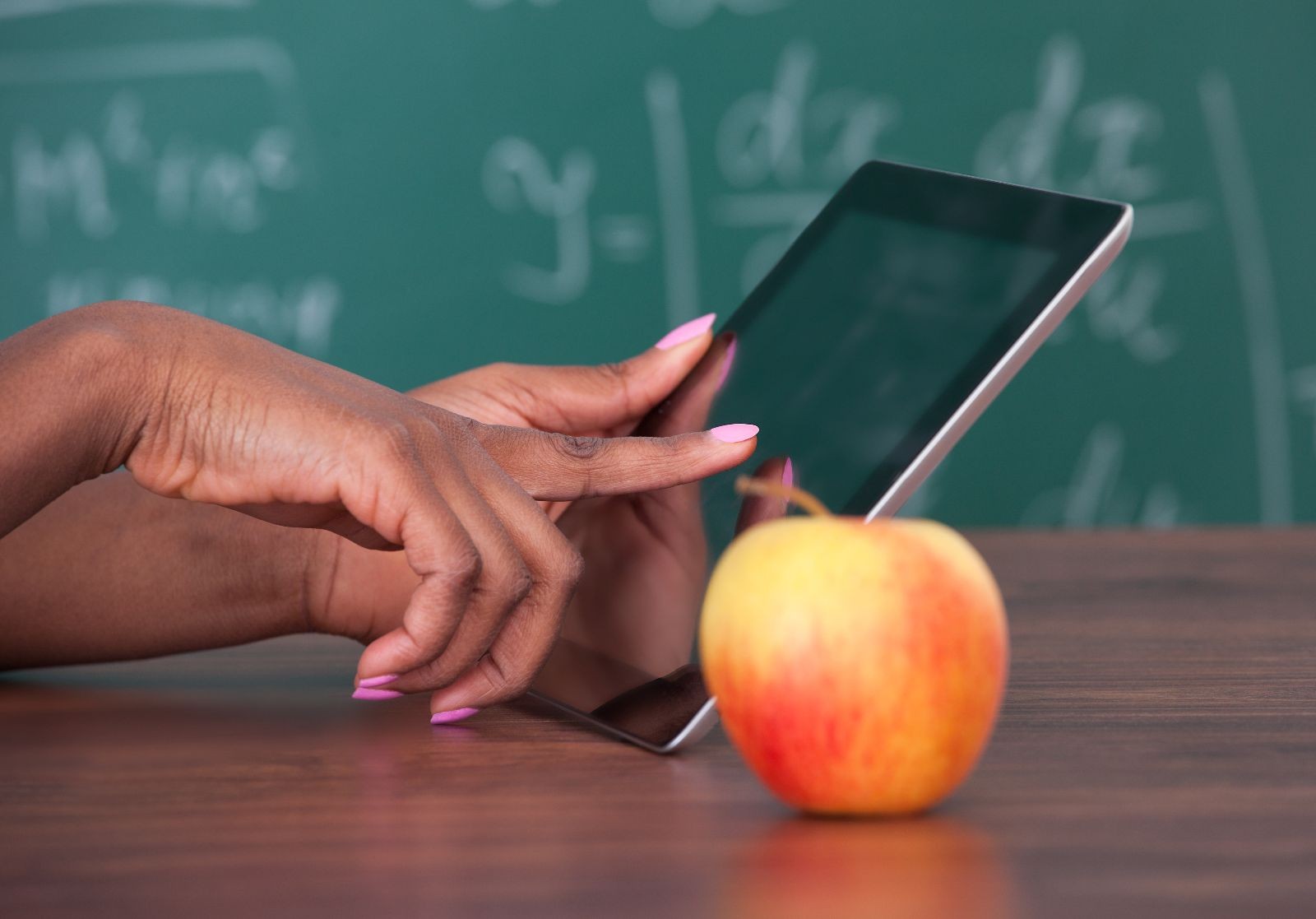 Profesora sostiene tableta digital en el escritorio con una manzana al fondo un pizarrón