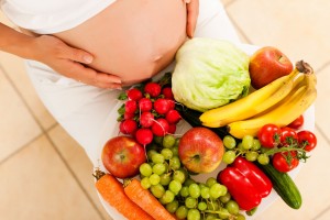 Mujer embarazada sentada con un plarón de frutas y verduras en sus piernas