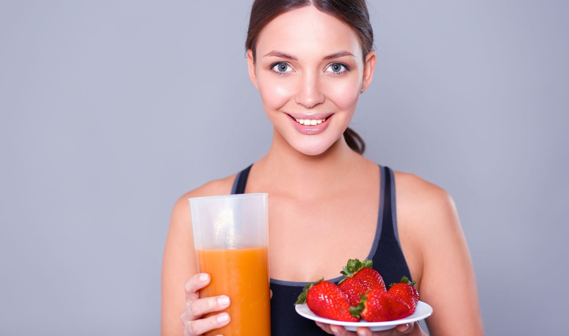 mujer joven sonriendo, sostiene con sus manos un vaso con jugo y un plato con fresas