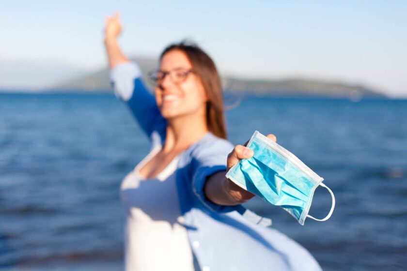 Mujer feliz quitarse la máscara médica en la playa del mar. Corona países libres. Final de cuarentena del coronavirus