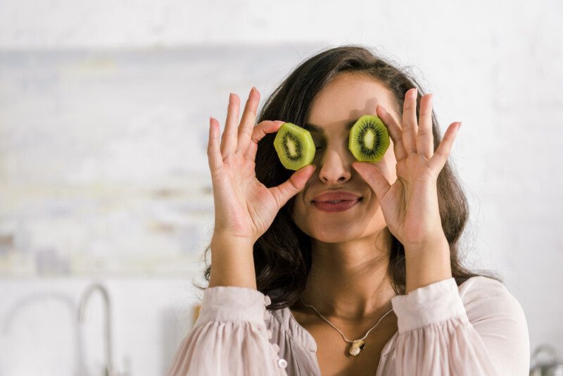 Mujer feliz sosteniendo mitades de kiwi mientras cubre los ojos