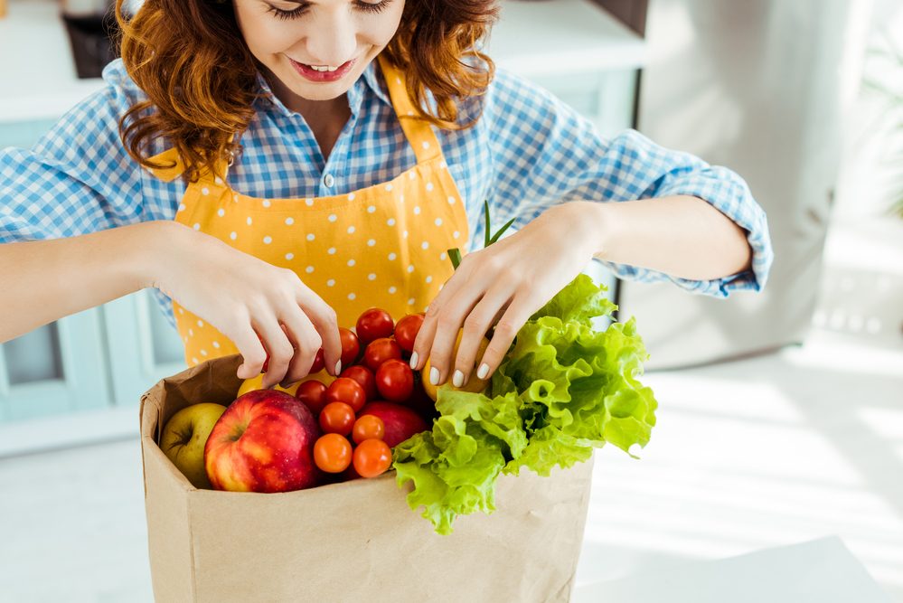 Mujer feliz en lunares delantal amarillo tomando frutas y verduras de la bolsa de papel