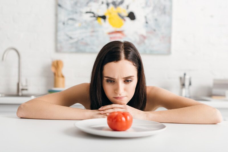 Chica pensativa mirando tomate maduro en la mesa de la cocina