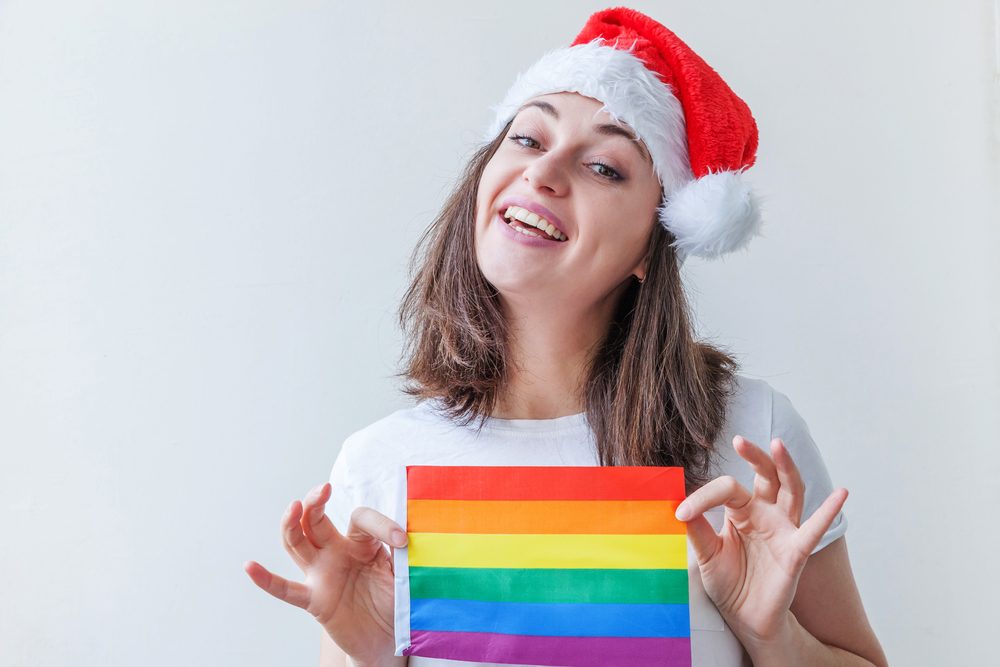 Hermosa chica lesbiana en el sombrero rojo de Santa Claus con la bandera del arco iris LGBT aislado en el fondo blanco mirando feliz y emocionado. Mujer joven retrato del Orgullo Gay. Feliz Navidad y Año Nuevo — Foto de Stock Hermosa chica lesbiana en el sombrero rojo de Santa Claus con la bandera de la diversidad