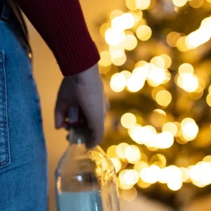 Person in front of the Christmas tree with a bottle of alcohol, drinking too much during Christmas time and New Year, woman